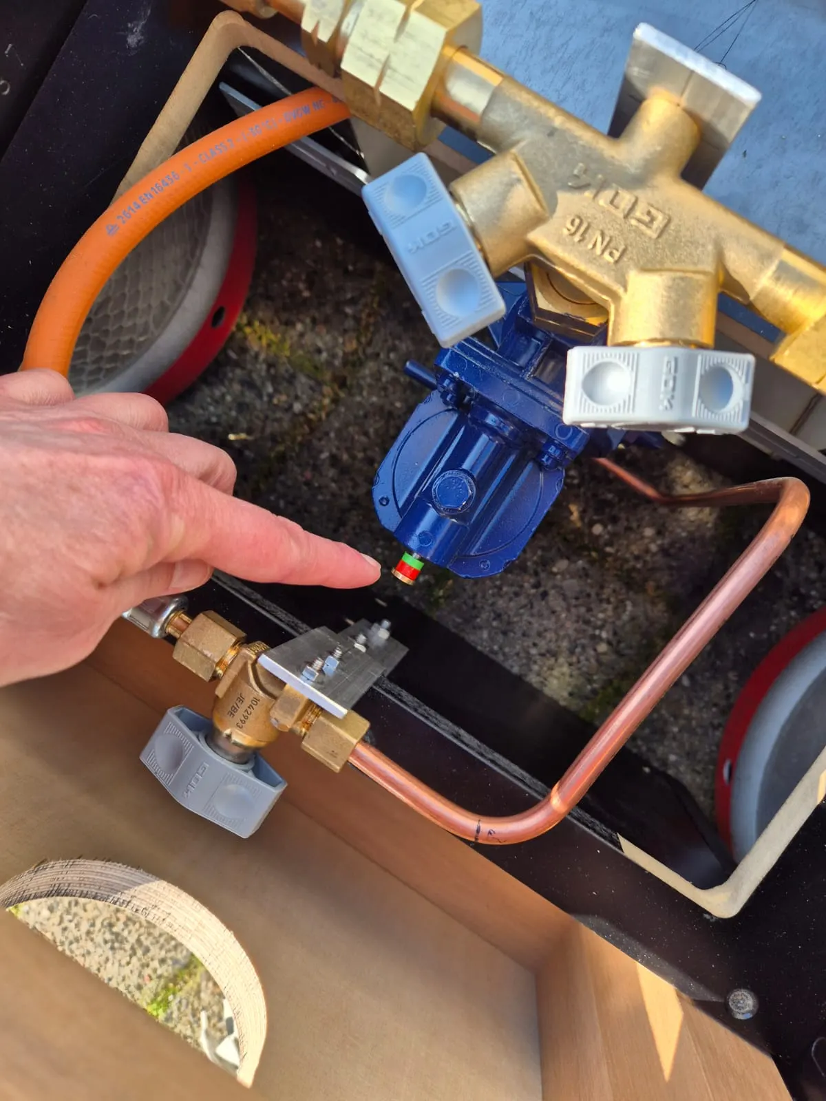 Eine Hand zeigt auf einen blauen Druckregler zwischen Messingarmaturen und Gasleitungen
